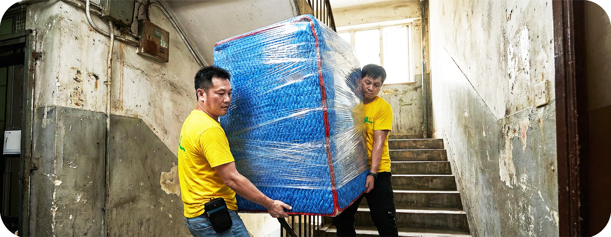 實惠搬屋團隊在唐樓狹窄樓梯間進行高難度搬運，兩位師傅合力將防撞包裹的重型傢俬搬運上樓。展示針對唐樓及村屋的樓梯搬運技術，強調樓梯費及推路費明碼實價，絕無隱藏收費。