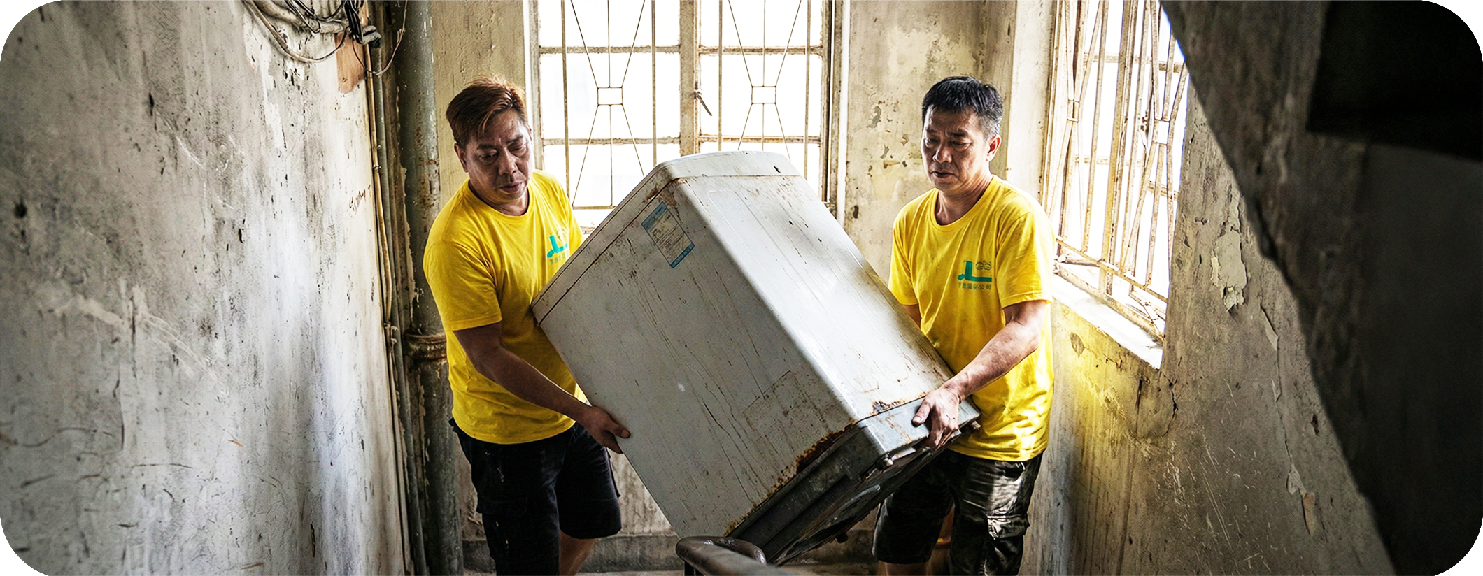 兩名實惠搬屋師傅合作在唐樓樓梯搬運舊洗衣機，提供合規的舊電器棄置及回收服務，解決無電梯大廈的重物搬運問題。